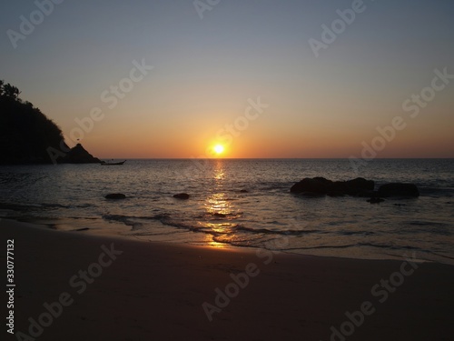 Reflection of sunset on the surface of sea water. Orange glow of sun. Stones and a mountain in the water on the background of an evening sundown. Waves, sand, deserted beach at twilight.