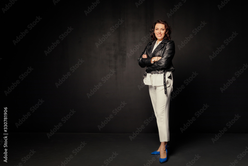 Smiling woman in a white suit and black leather jacket standing and looking at the camera on a black background. Free space