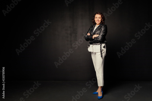 Smiling woman in a white suit and black leather jacket standing and looking at the camera on a black background. Free space