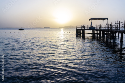 View of the calm sea at dawn with pink clouds on the horizon and wooden pier and ship in the distance