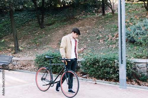 Young hispanic man using mobile and carrying a bike while walking outdoors