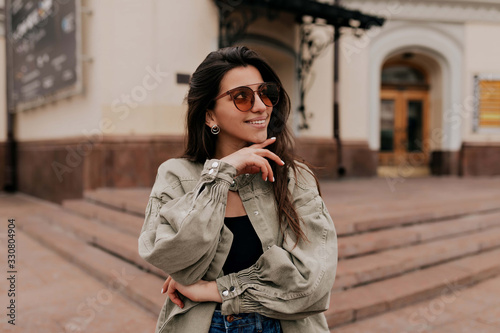 Obraz na płótnie Close-up portrait of laughing brunette girl walking on city background. Happy woman in sunglasses and dress walking outdoors. Image of beautiful stylish woman on street on spring day w salonie