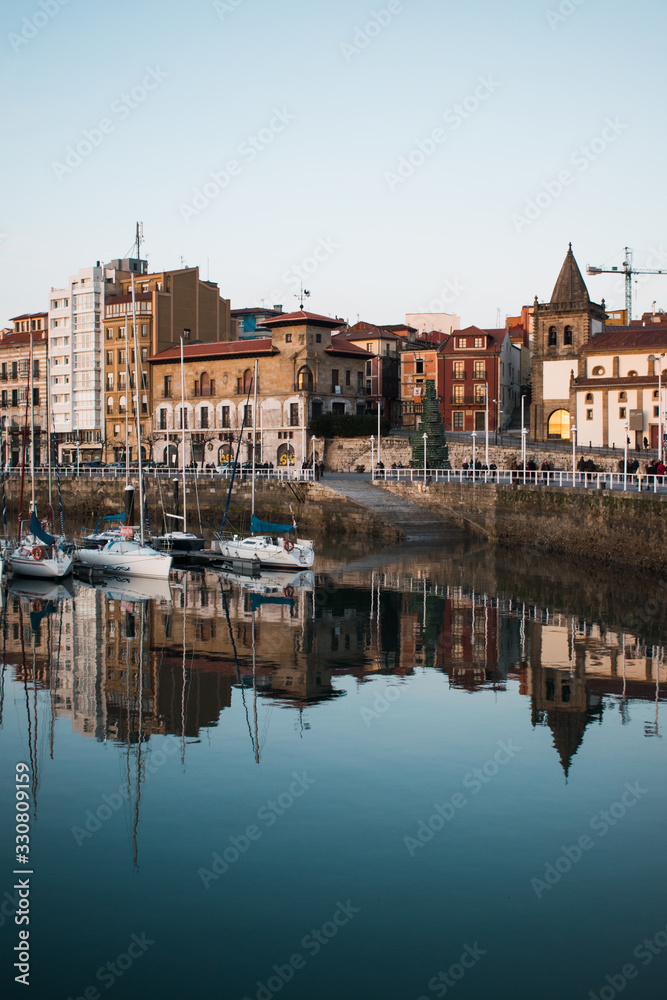 Barrio de Cimadevilla y barcos de vela reflejados en las aguas del puerto en una tarde tranquila