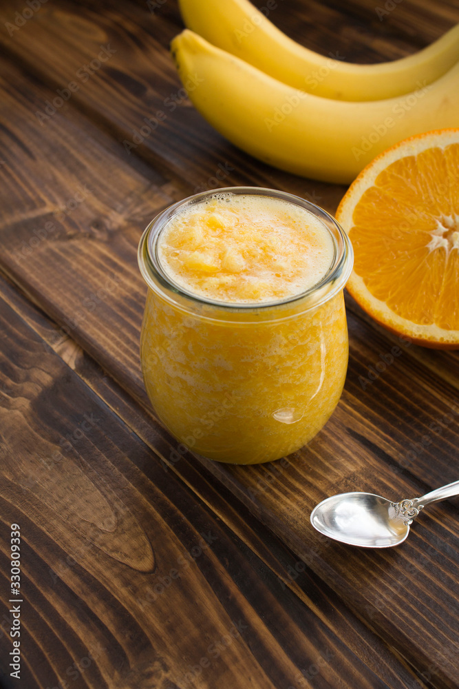 Smoothies or puree with orange and banana in the small glass jar on the brown wooden background. Location vertical. Smoothies or puree with orange and banana in the small glass jar on the brown wooden background. Location vertical.