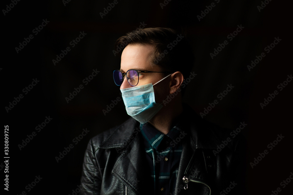 Closeup view of  man with glasses and protective surgical mask dressed in black clothes on a black background. Pandemic covid 19. Concept for the prevention of viral diseases. Studio photo.