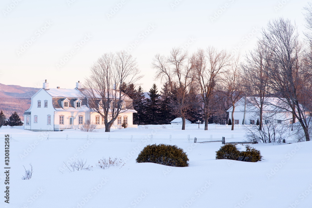 Winter landscape with beautiful patrimonial double white house with Mansard roof and large wooden barn set in snowy field, St. Pierre, Island of Orleans, Quebec, Canada
