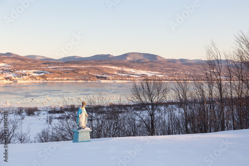 Old traditional roadside Virgin May statue seen in snowy field with the St. Lawrence River and the Laurentian mountains in the background, St. Pierre, Island of Orleans, Quebec, Canada