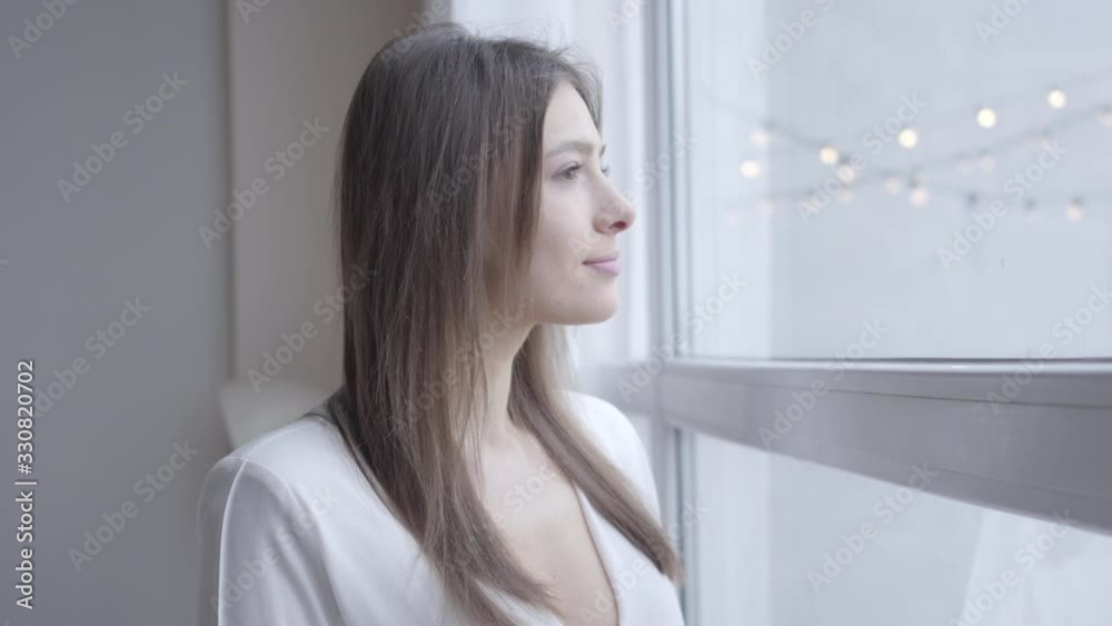 Close-up of thoughtful brunette Caucasian woman with brown eyes looking out the window at home. Cute girl waking up in the morning on weekends. Lifestyle, joy, leisure.