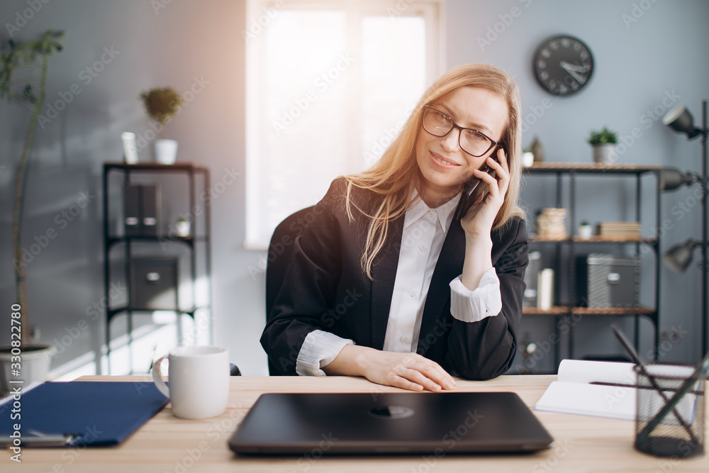 Mature business woman with blond hair wearing eyeglasses and black suit having business conversation on smartphone while sitting at workplace. Concept of working process and career Mature business woman with blond hair wearing eyeglasses and black suit having business conversation on smartphone while sitting at workplace. Concept of working process and career