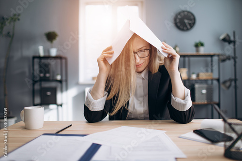 Tired mature lady in formal clothing stressing while working with paper documents at modern office. Female manger in eyeglasses sitting at table and doing paper work.