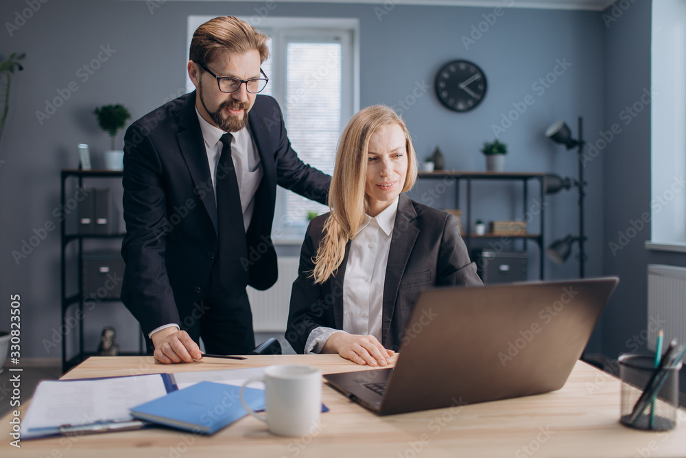 Mature lady with blond hair sitting at working place and looking at computer screen while male business partner standing near and consulting her about new project. Concept of teamwork. Mature lady with blond hair sitting at working place and looking at computer screen while male business partner standing near and consulting her about new project. Concept of teamwork.