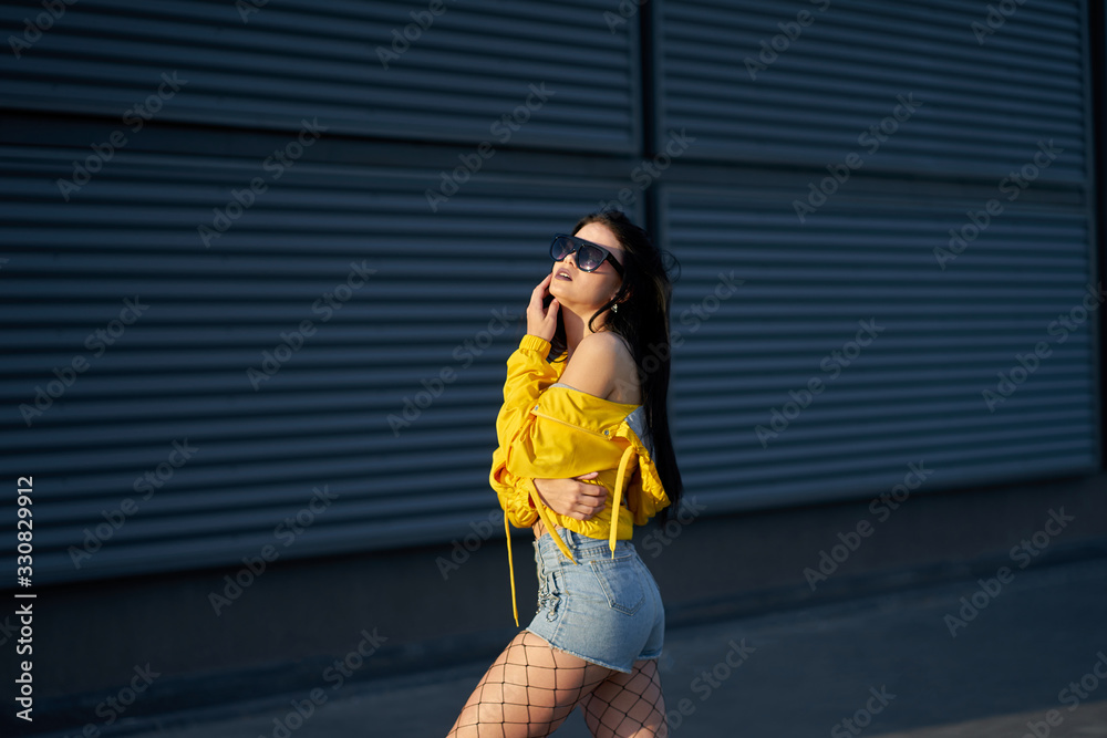 Fototapeta premium Fashion. Stylish hipster brunette girl dressed youth yellow jacket, denim shorts and fashionable sunglasses posing on grey metal urban wall background.
