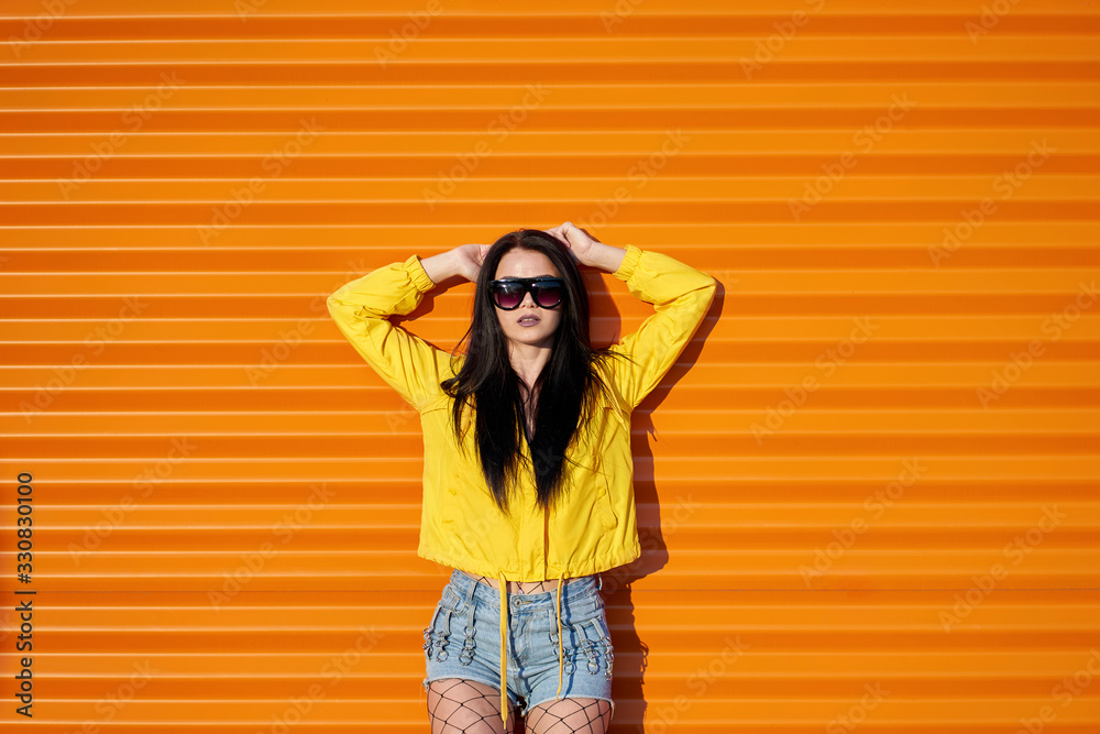 Beautiful sexy young stylish girl with long brunette hair wearing trendy yellow jacket, denim shorts and fashionable sunglasses posing on orange urban wall background.
