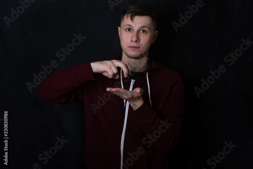 Studio portrait of a young man in a red sweatshirt against black background. A deaf-mute guy shows a "jump" gesture with his fingers. Sign language.