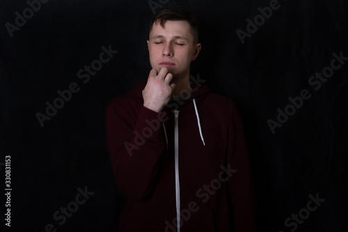 Studio portrait of a young man in a red sweatshirt against black background. A deaf and dumb guy shows a “sleep” gesture with his fingers. Eyes closed. Shows a sleeping person. Sign language.