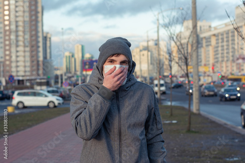 portrait of a guy in the city. on the face a medical white mask to protect against viruses. Coronavirus is transmitted by airborne droplets. He covers his nose and mouth with his hand. © Anton
