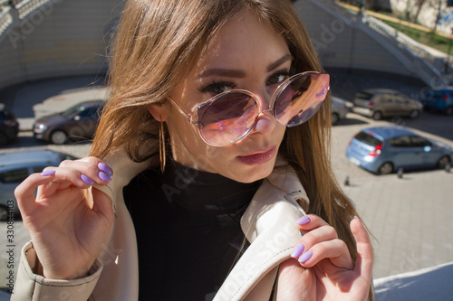 Street Fashion young attractive girl in a beige jacket, black turtleneck, black shiny pants and beige shoes against the background of urban locations