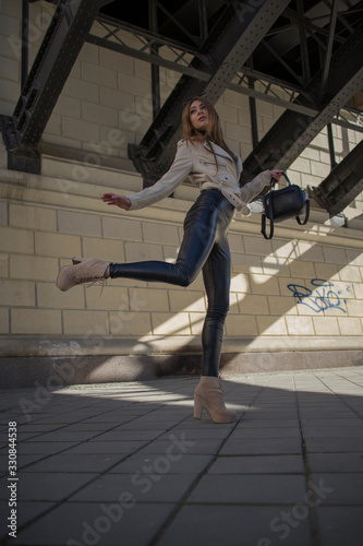 Street Fashion young attractive girl in a beige jacket, black turtleneck, black shiny pants and beige shoes against the background of urban locations © Ruslan