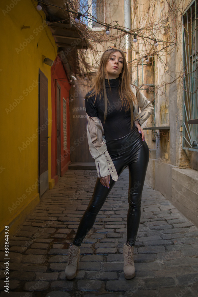 Street Fashion young attractive girl in a beige jacket, black turtleneck, black shiny pants and beige shoes against the background of urban locations