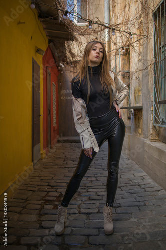 Street Fashion young attractive girl in a beige jacket, black turtleneck, black shiny pants and beige shoes against the background of urban locations
