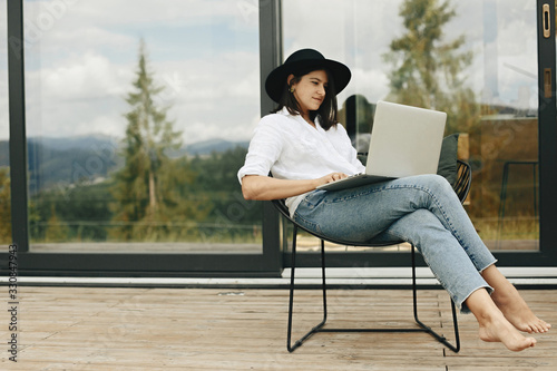 Fotobehang Stylish hipster girl sitting with laptop on terrace at big window
