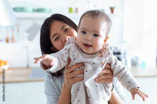 Happy young mother with her baby daughter looking at camera in the kitchen at home.