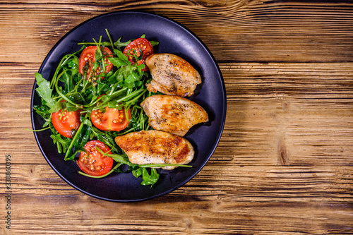 Roasted chicken breasts and salad with arugula and cherry tomatoes in a black plate. Top view