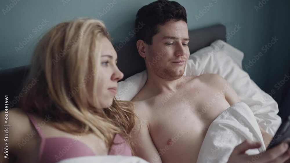 Man who is addicted to the phone refusing his girlfriend in inntimacy. Woman frustrated because of her boyfriend doesn't pay attantion to her in bed. Millenial problems, fabbing, sex concept. Man who is addicted to the phone refusing his girlfriend in inntimacy. Woman frustrated because of her boyfriend doesn't pay attantion to her in bed. Millenial problems, fabbing, sex concept.