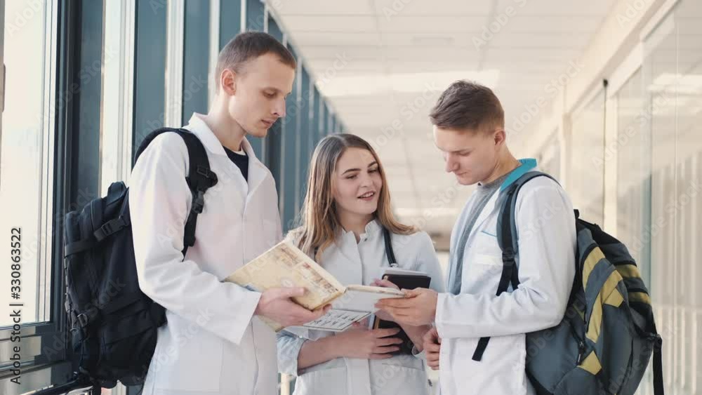 Medical students with books and bags are discussing something in a modern hall. There are two European boys and a girl in robes. Medical students with books and bags are discussing something in a modern hall. There are two European boys and a girl in robes.