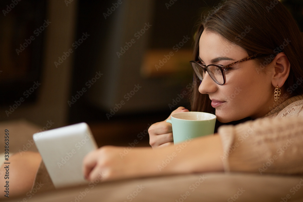 Woman sitting on the carpet near the sofa using a tablet, drinking coffee from a cup. Online education concept.