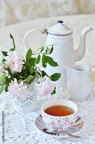 A tea party in the style of Shabby Chic. tea in the beautiful tea steam with a pattern "rose". Delicate pink roses in a vase, white kettle in retro style. On a white lace tablecloth. Soft focus.