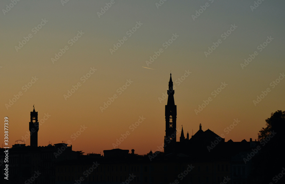 Church towers and the tower of the Palazzo Vecchio, Florence, are seen in silhouette against the colours of sunset sunset. The sky is clear, with a vapour trail left by a plane.