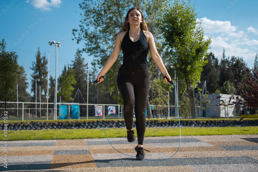 Mujer joven fitness con ropa oscura brinca la cuerda en un parque durante un día verde soleado Mujer joven fitness con ropa oscura brinca la cuerda en un parque durante un día verde soleado