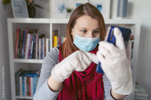 Young woman wearing protective gloves on hands and mask on face working from home or at office work by the table laptop mobile phone video call preventing virus spread epidemic quarantine prevention