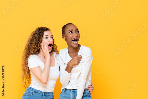 Excited shocked interracial woman friends looking aside isolated on yellow studio background