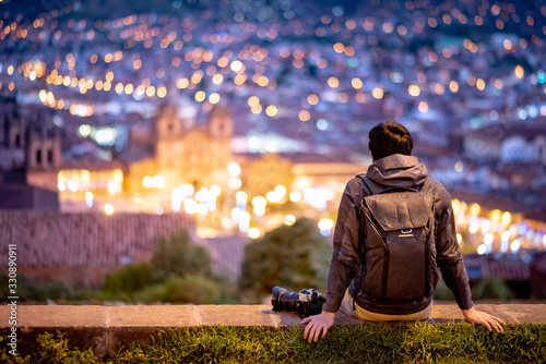 Asian man traveler and photograpaher sitting on viewpoint looking at illuminated cusco city at night. Cusco (Cuzco) is a city in southeastern Peru, near the Urubamba Valley of the Andes mountain range