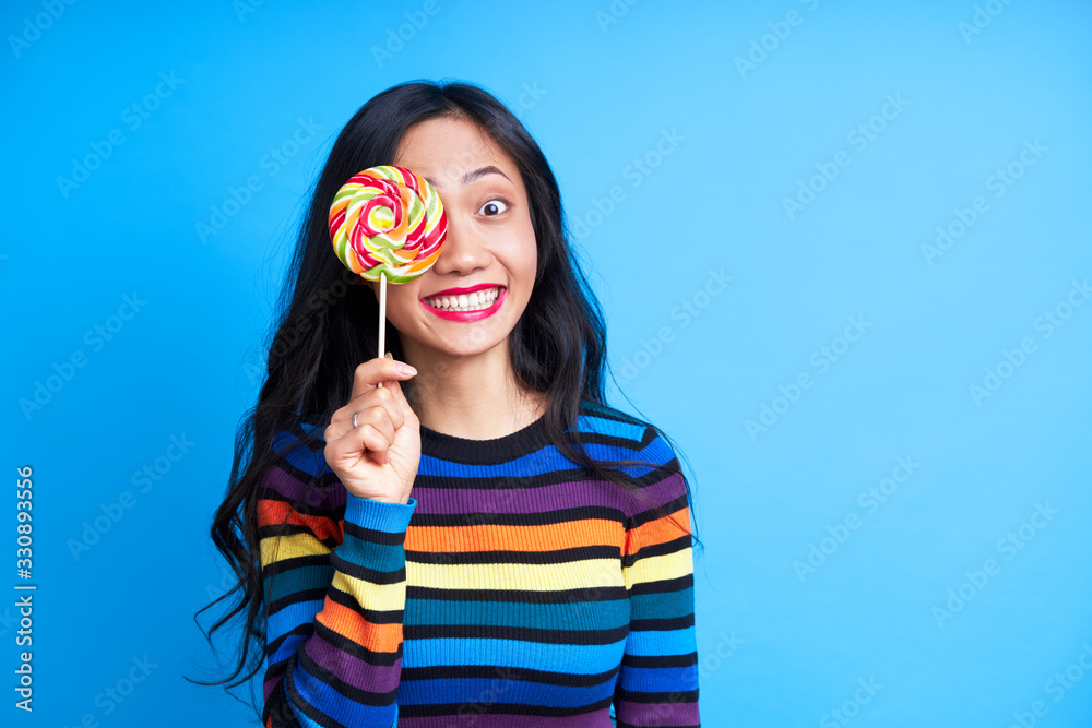 Happy beautiful woman covers her eye with colorful lollipop isolated on blue background. Happy beautiful woman covers her eye with colorful lollipop isolated on blue background.