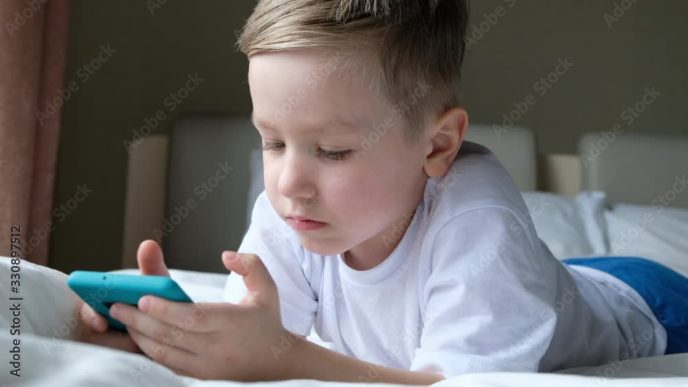 Cute little boy toddler playing mobile game, lying on a bed and holding smartphone Cute little boy toddler playing mobile game, lying on a bed and holding smartphone