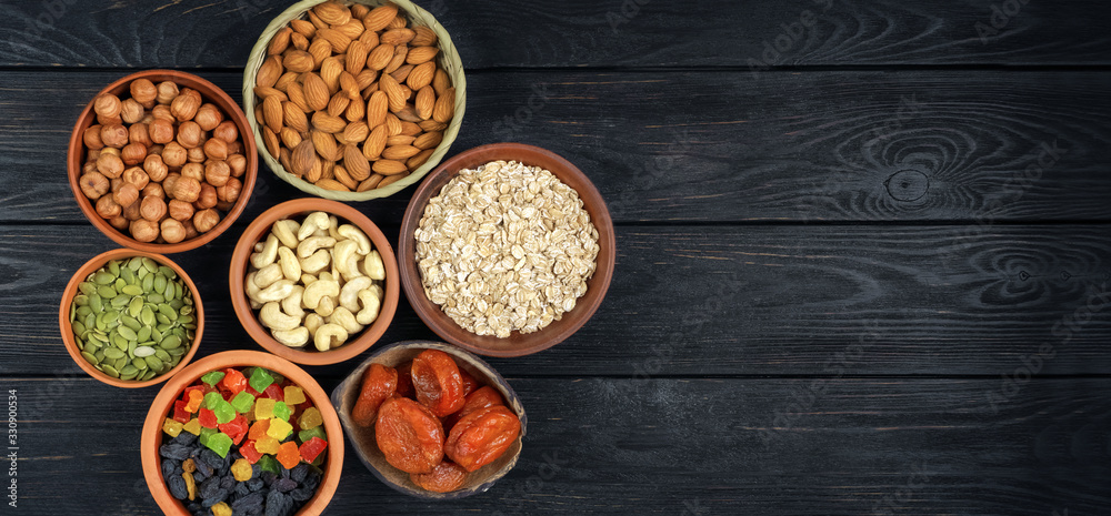 Granola. Oatmeal, barley flakes. Nuts: hazelnuts, almonds, cashews. Dried fruits: raisins, dates, dried apricots. In clay cups on a black, wooden background. Top view. Flat lay. Granola. Oatmeal, barley flakes. Nuts: hazelnuts, almonds, cashews. Dried fruits: raisins, dates, dried apricots. In clay cups on a black, wooden background. Top view. Flat lay.