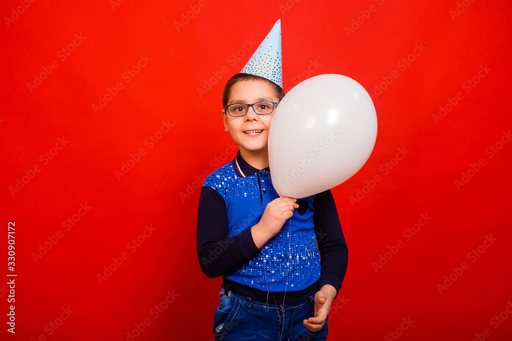 A cheerful boy in a festive cap with a balloon in his hands stands on a red background. A cheerful boy in a festive cap with a balloon in his hands stands on a red background.