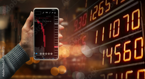 A hand holding a black smartphone showing stock market candlestick trend on blurred-out stock market data display panel, Business and finance concept.