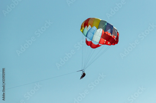 With a parachute for parasailing a couple flies through the air with blue sky in the background. Happy couple hovers in the air on a multi-colored parachute and admires the scenery from a bird 's eye