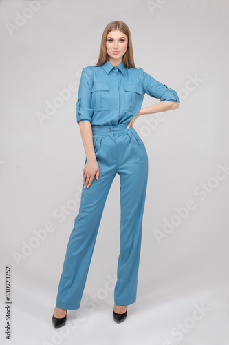 Full length stock photo of a gorgeous slim and tall model with make up and fair hair wearing elegant blue shirt and trousers with black heels. She is posing over white background with hand on waist.