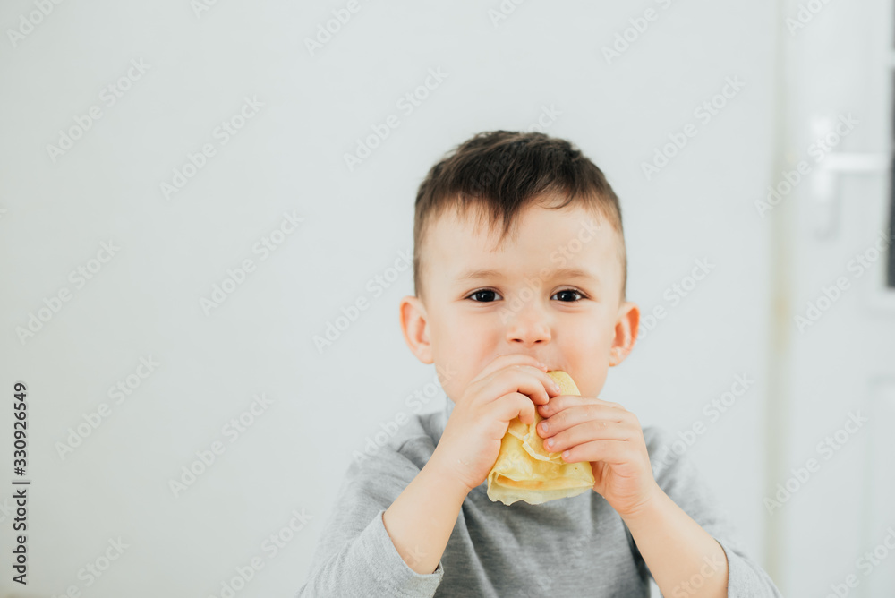 Cute boy eating delicious pancakes sitting at a table on a light background Cute boy eating delicious pancakes sitting at a table on a light background