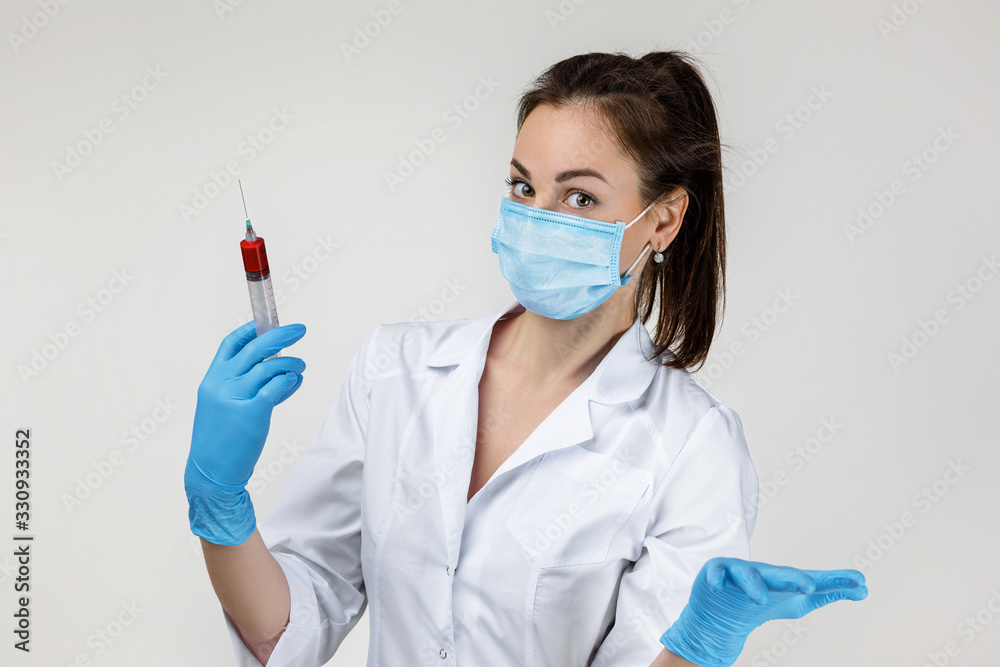 Young woman doctor in white uniform, mask and rubber gloves holding syringe on gray background. Young woman doctor in white uniform, mask and rubber gloves holding syringe on gray background.