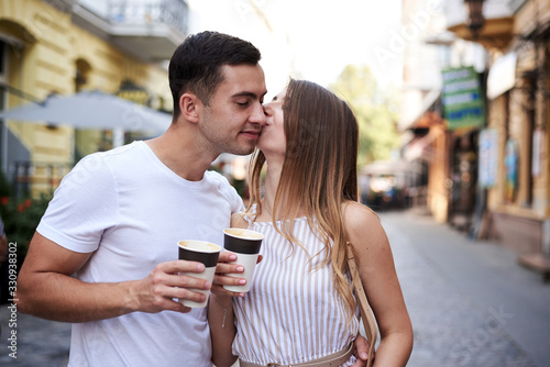 Romantic couple on date drinking coffee cappuccino in paper cups on street . Young blond woman  walking with brunette man with yummy coffee drink in summer in morning. Nutritious breakfast  snack.