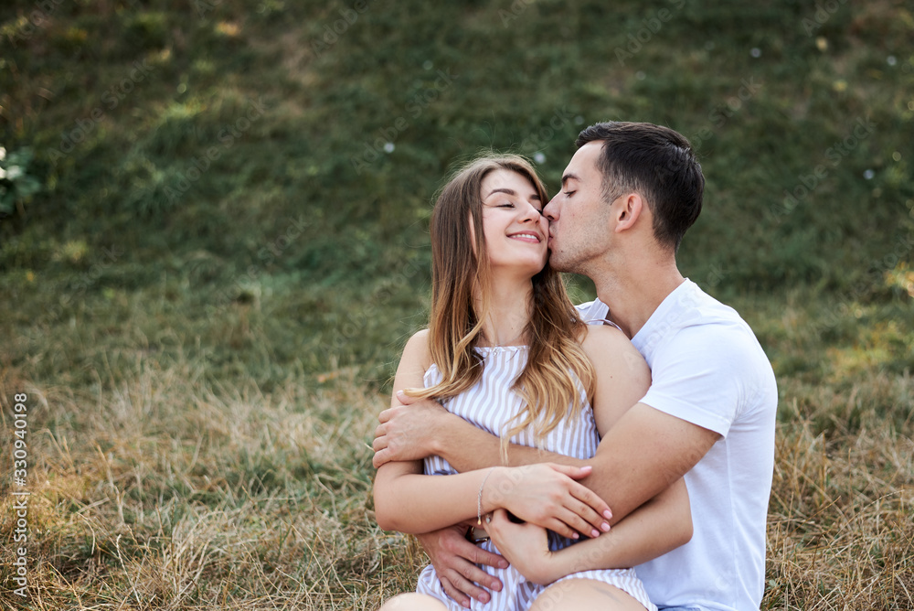 Young couple in love sitting on green yellow grass lawn hugging embracing kissing. Blond woman wearing stripy short overall and brunette man in white t-shirt blue shorts on romantic date. Relationship Young couple in love sitting on green yellow grass lawn hugging embracing kissing. Blond woman wearing stripy short overall and brunette man in white t-shirt blue shorts on romantic date. Relationship