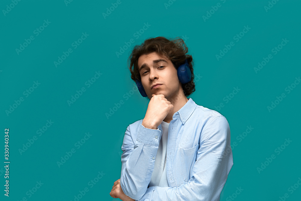 Portrait of a young beautiful man wearing white t-shirt and blue shirt in blue headphones thought holding a fist at his chin and looking at the camera over blue background