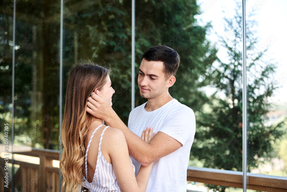 Naklejka premium Young couple in love hugging by wooden railing, glass window and green trees on terrace. Blond woman, wearing stripy short overall and brunette man in white t-shirt and blue shorts on romantic date.