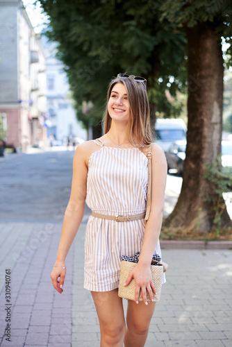 Young pretty blond woman, wearing white stripy short overall, walking on small alley avenue walkway in summer. Three-quarter portrait of slim girl posing for picture in resort. Vacation leisure time